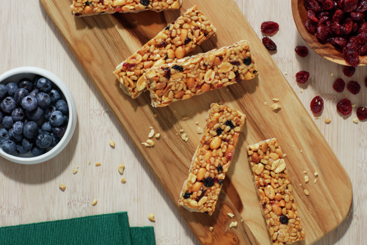 Five Nature Valley bars randomly arranged on a cutting board with bowls of blueberries and craisins