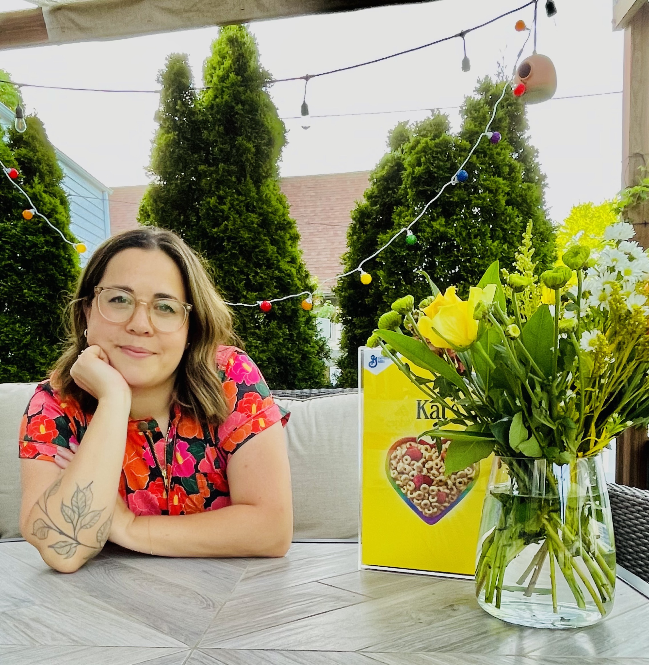 Candid photo of Kat Ricker employee and President of our General Mills' Betty's Family, LGBTQ+ Employee Network, sitting at a table with a personalized Cheerios cereal box and vase of yellow flowers.